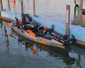 A sit-on-top fishing kayak with orange accents and a mounted trolling motor docked at a wooden pier in a marina.