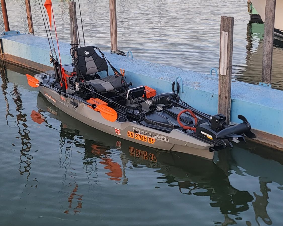 A sit-on-top fishing kayak with orange accents and a mounted trolling motor docked at a wooden pier in a marina.