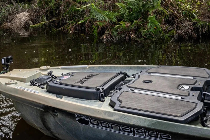 A person in khaki pants stands on the deck of a small fishing boat on shallow water, with dense green shoreline in the background. The boat has a dark gray seat and black storage compartments.